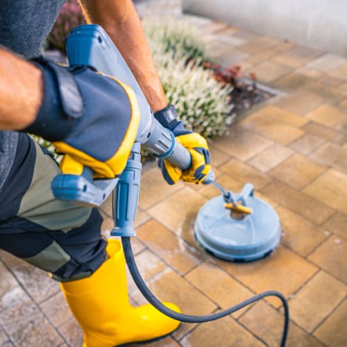 Person Using A Pressure Washer On A Patio In A Residential Backyard During Daytime